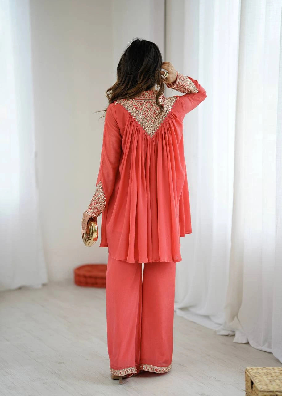 Woman wearing a coral pink traditional outfit with gold embroidery in a minimalistic setting.