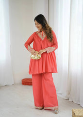 Woman in a coral outfit with gold details holding a gold clutch against a white curtain background