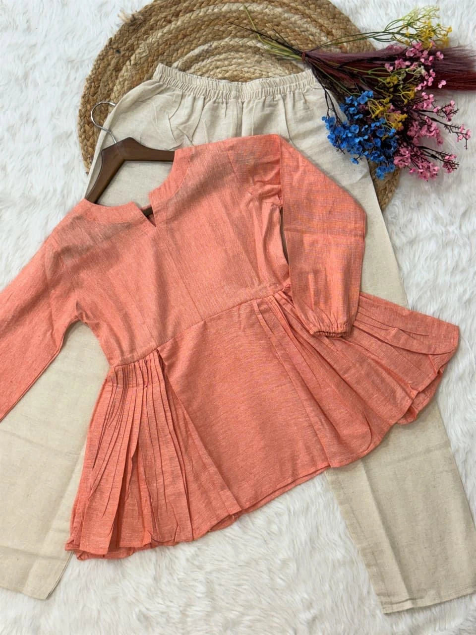 Coral long-sleeve top on a beige hanger with flowers and a hat in the background