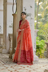 Woman in a red saree standing in an outdoor setting with greenery.