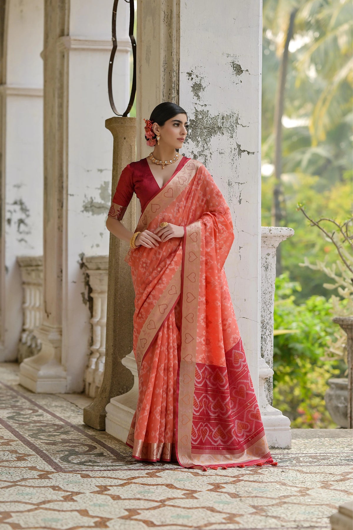 Woman in a red and pink saree standing in an outdoor setting with greenery.