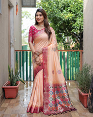 Woman in a peach and pink saree standing outdoors with plants and a building in the background