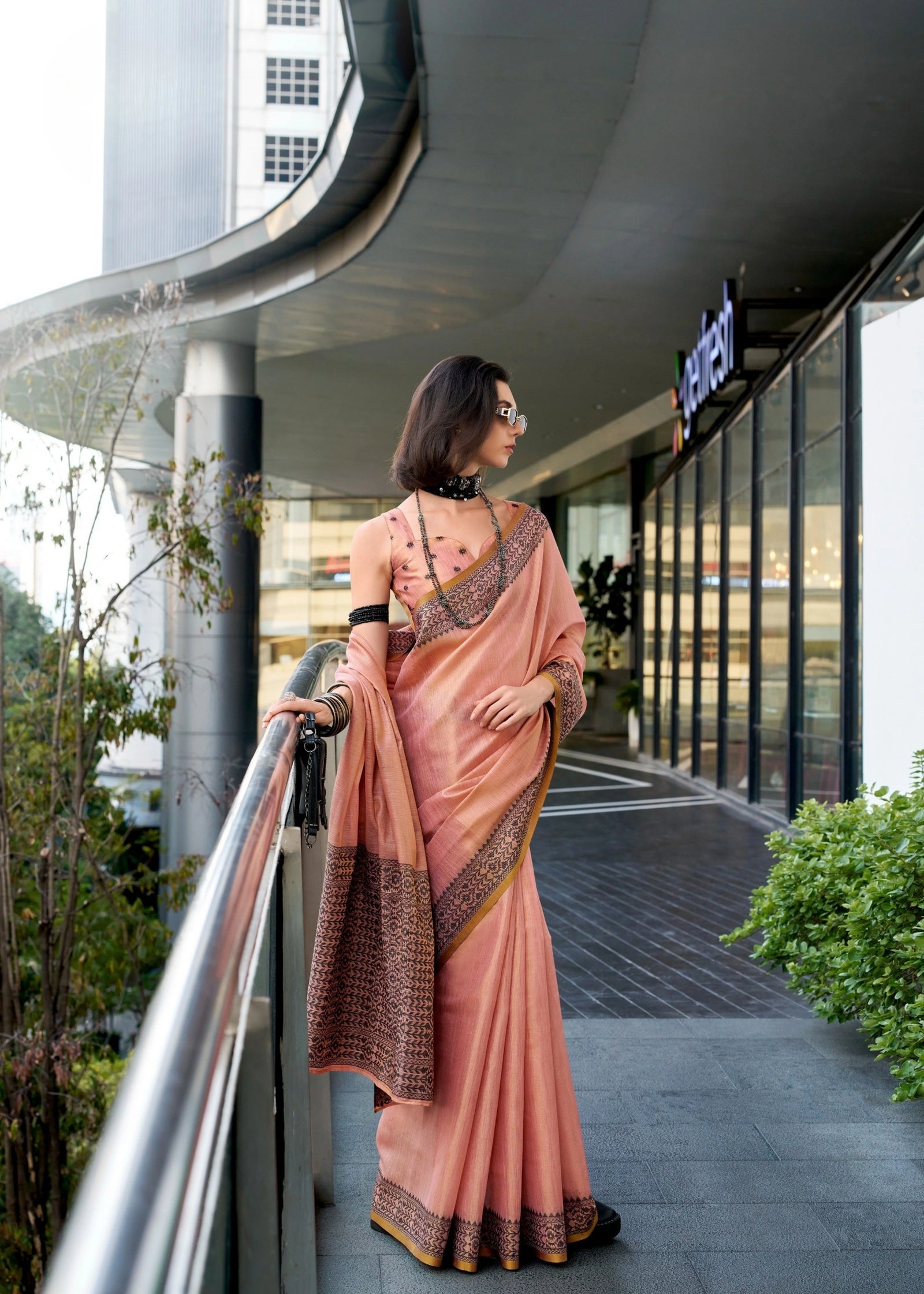 Woman in a peach saree standing on a balcony with modern architecture in the background