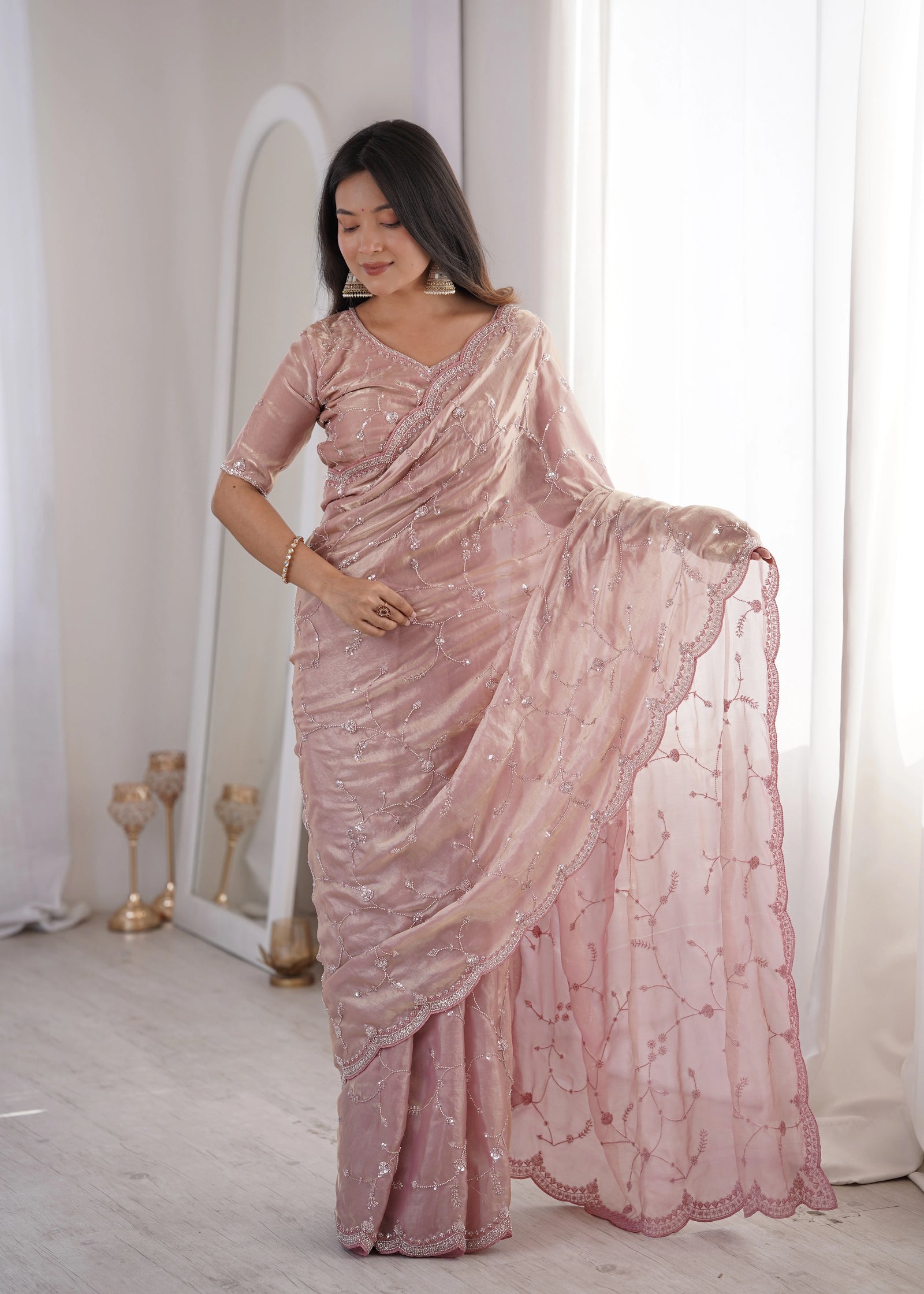 Woman wearing a pink saree in a well-lit room with white curtains.