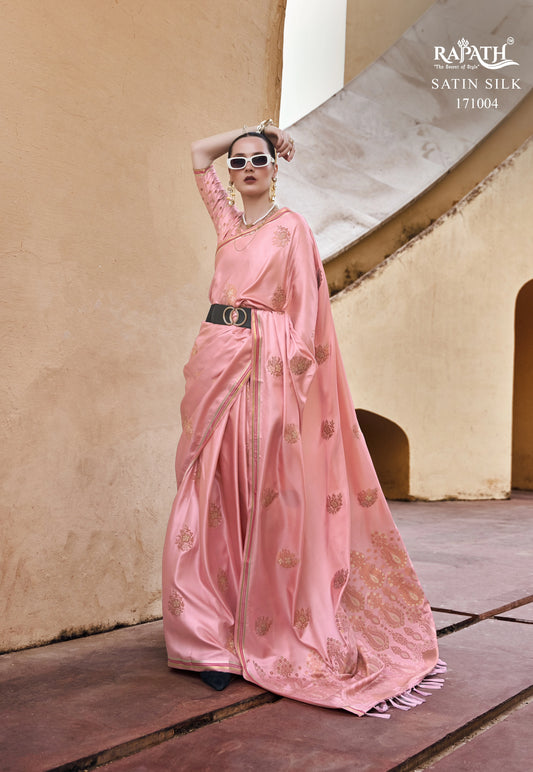 Woman in a pink saree with gold patterns standing against a beige wall.