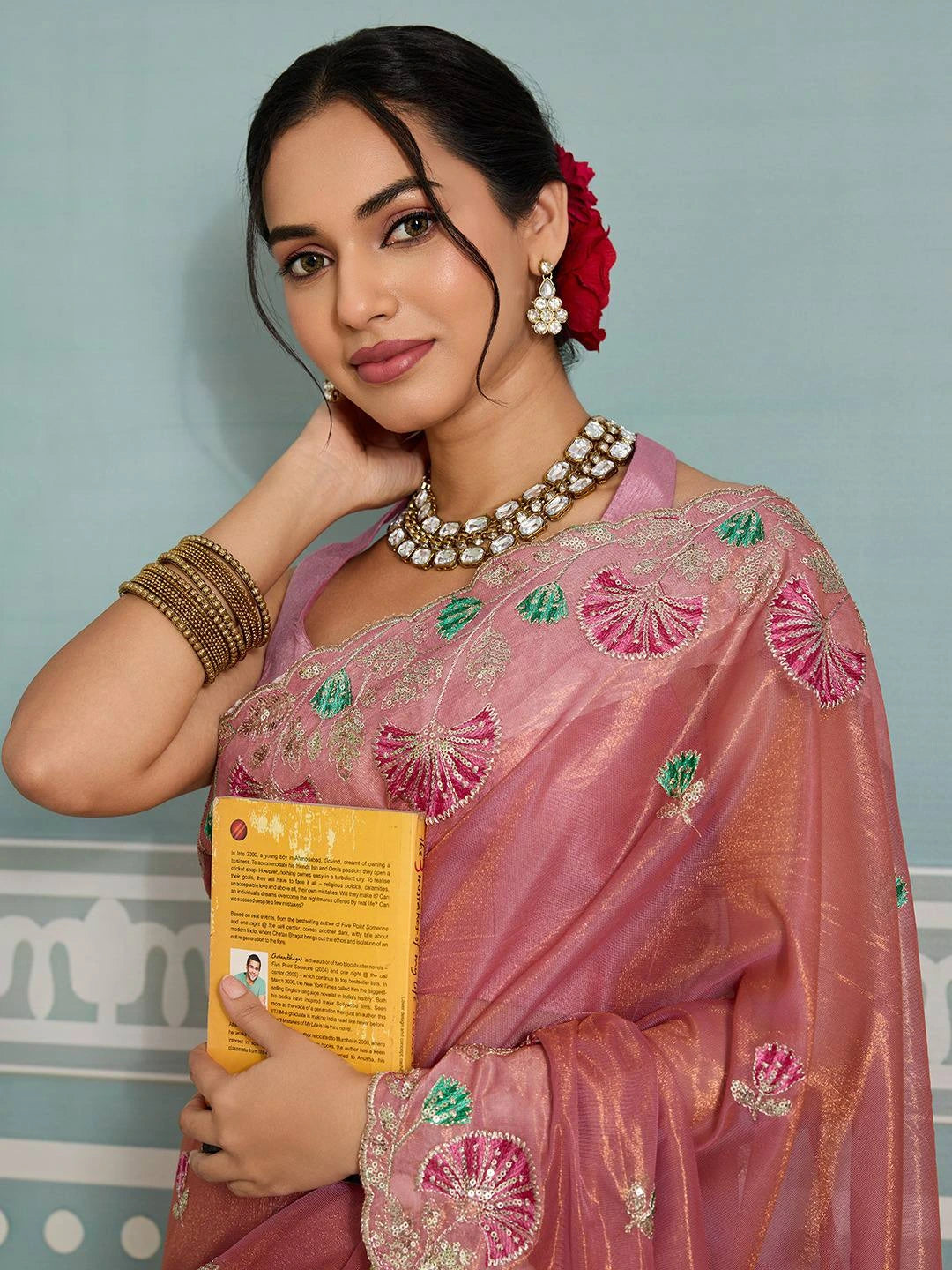 Woman in traditional pink saree with floral decorations, holding a yellow card against a light blue wall.