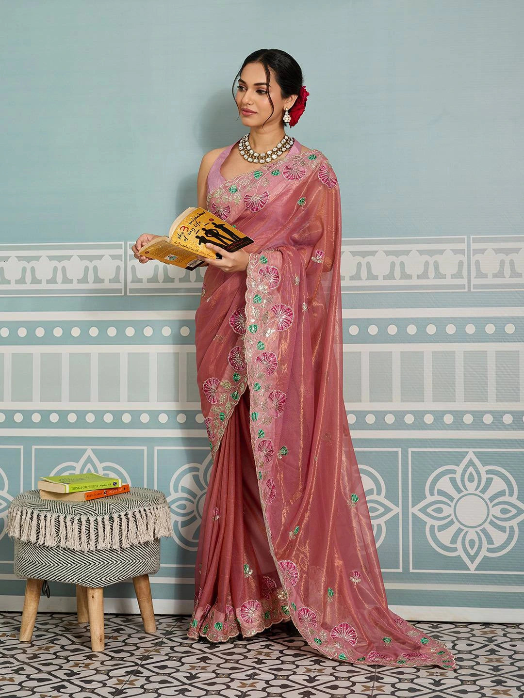 Woman in a pink saree with floral patterns standing in a room with decorative wall and stool.