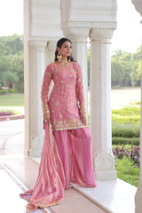 Woman in a pink traditional outfit standing in front of white architectural columns with greenery in the background.