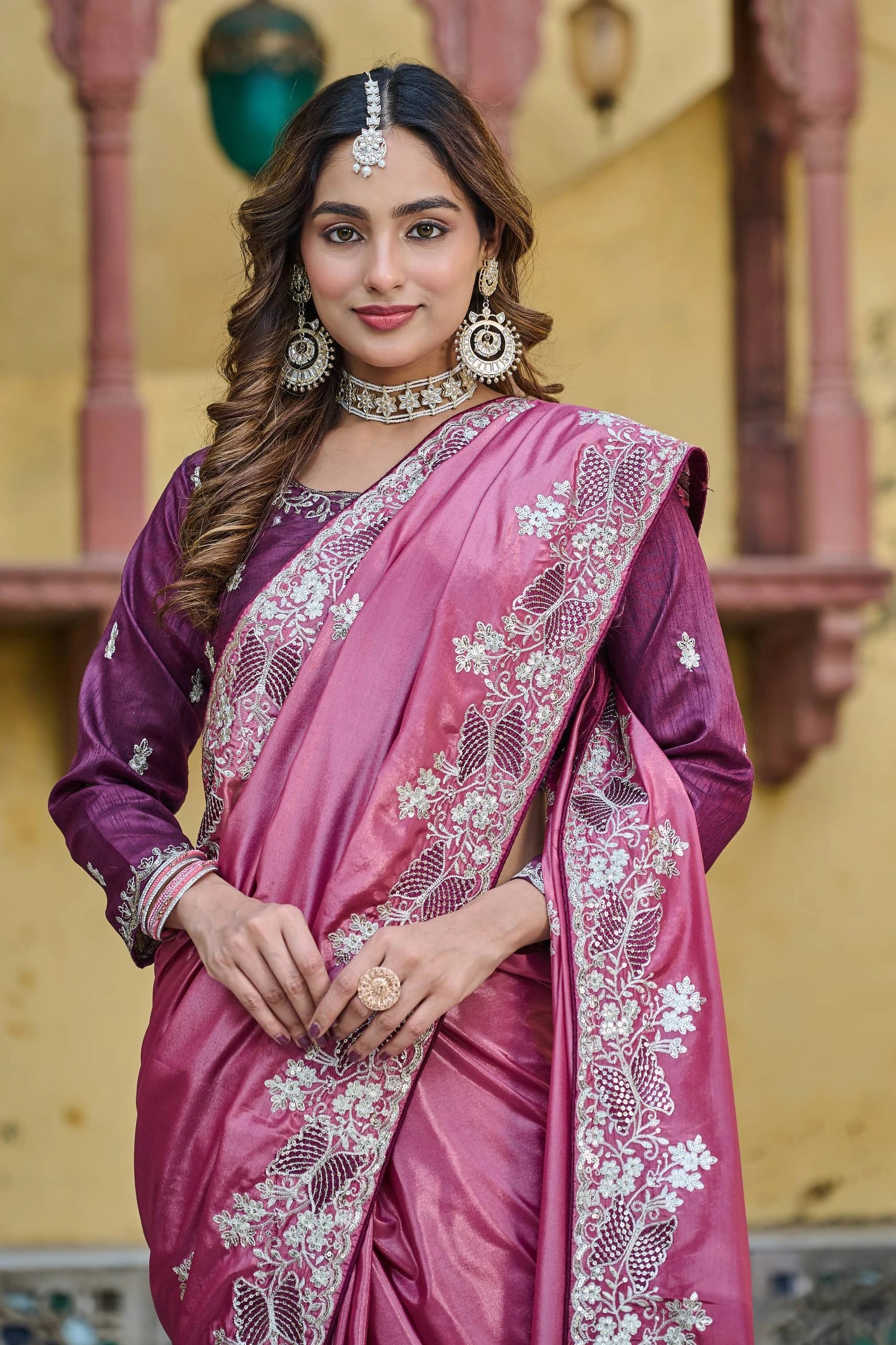 Woman wearing a pink saree with intricate designs and jewelry in an indoor setting.