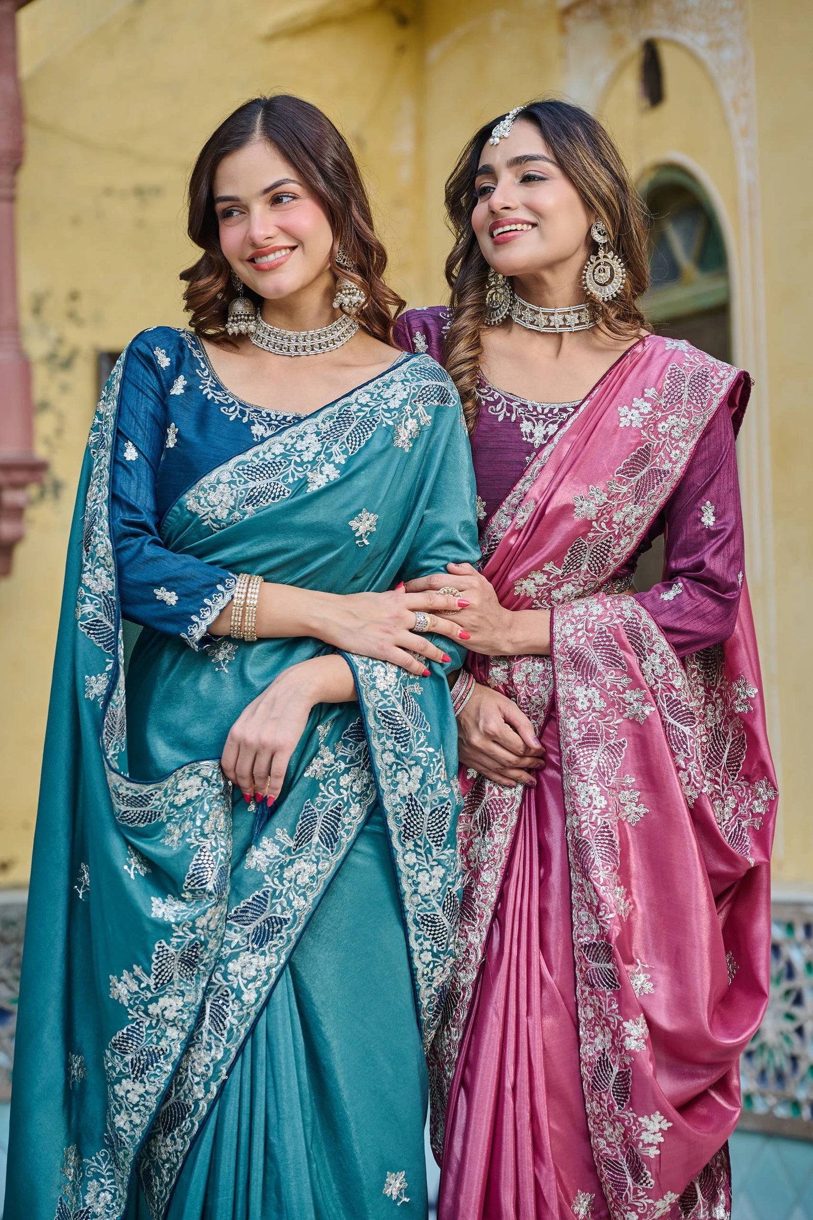 Two women in traditional sarees standing together with a architectural background