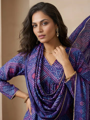 Woman wearing a purple patterned outfit with gold jewelry against a beige background