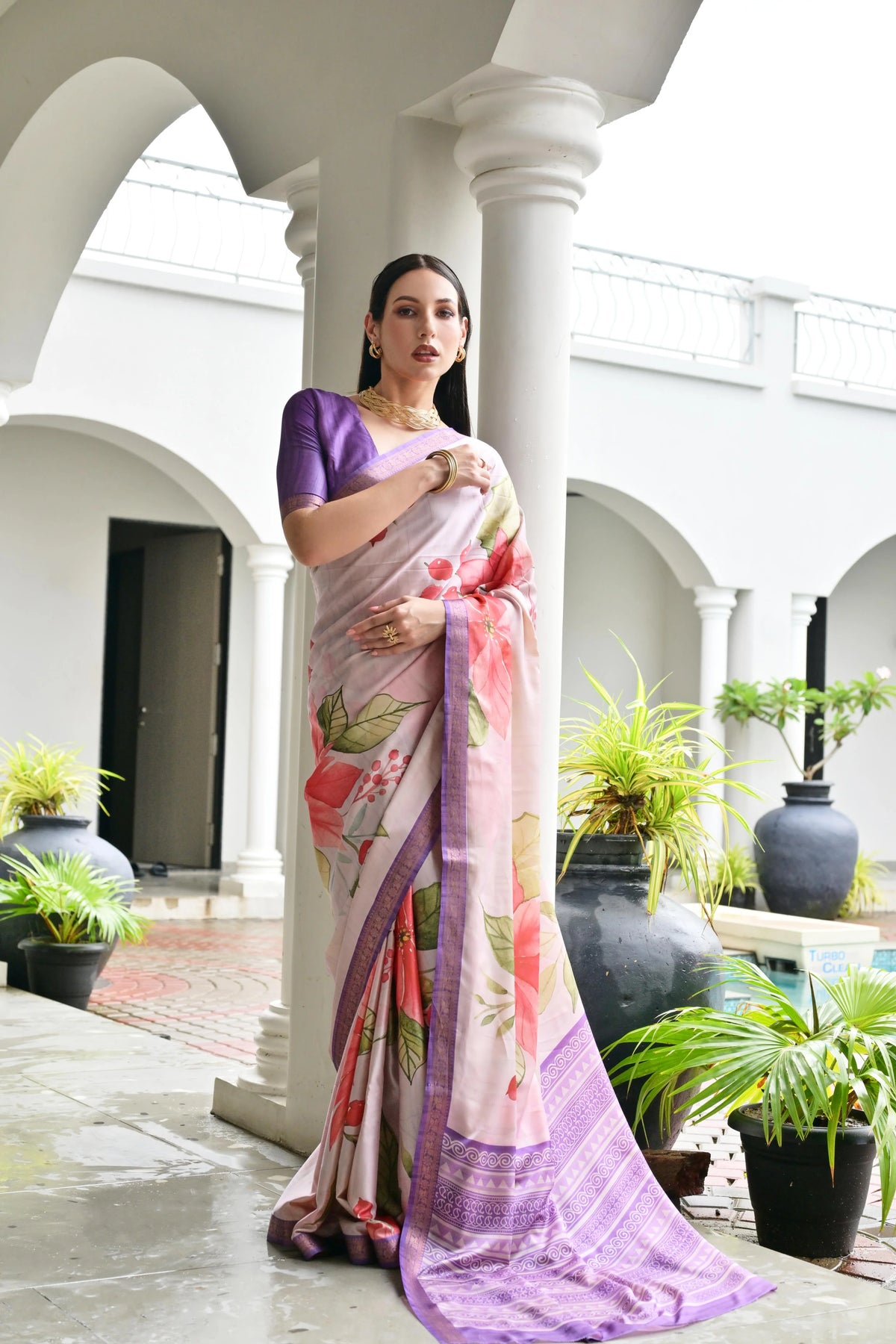 Woman in a floral saree standing in an outdoor setting with plants and architectural columns.