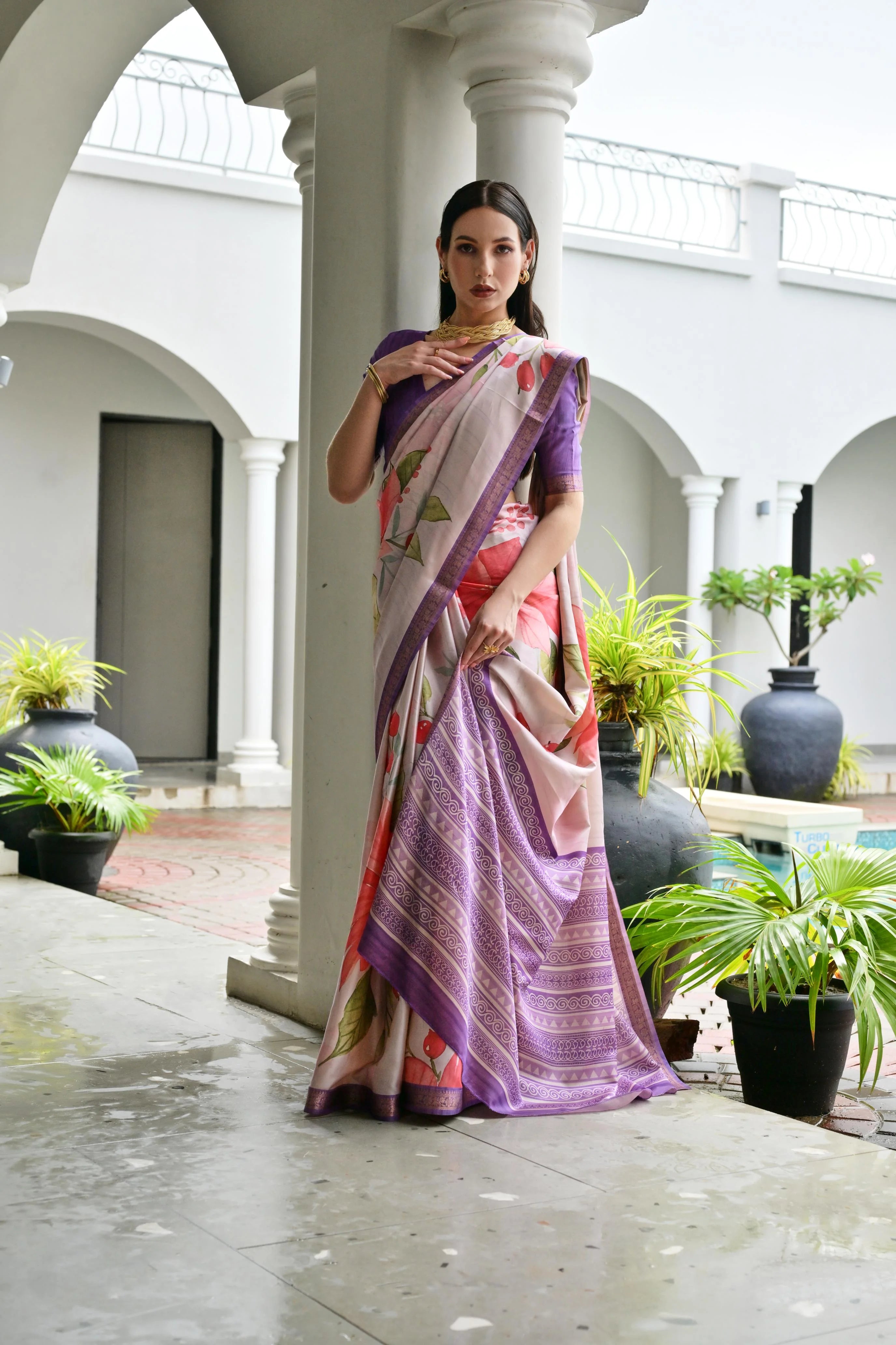 Woman in a colorful saree standing in an outdoor setting with columns and plants.