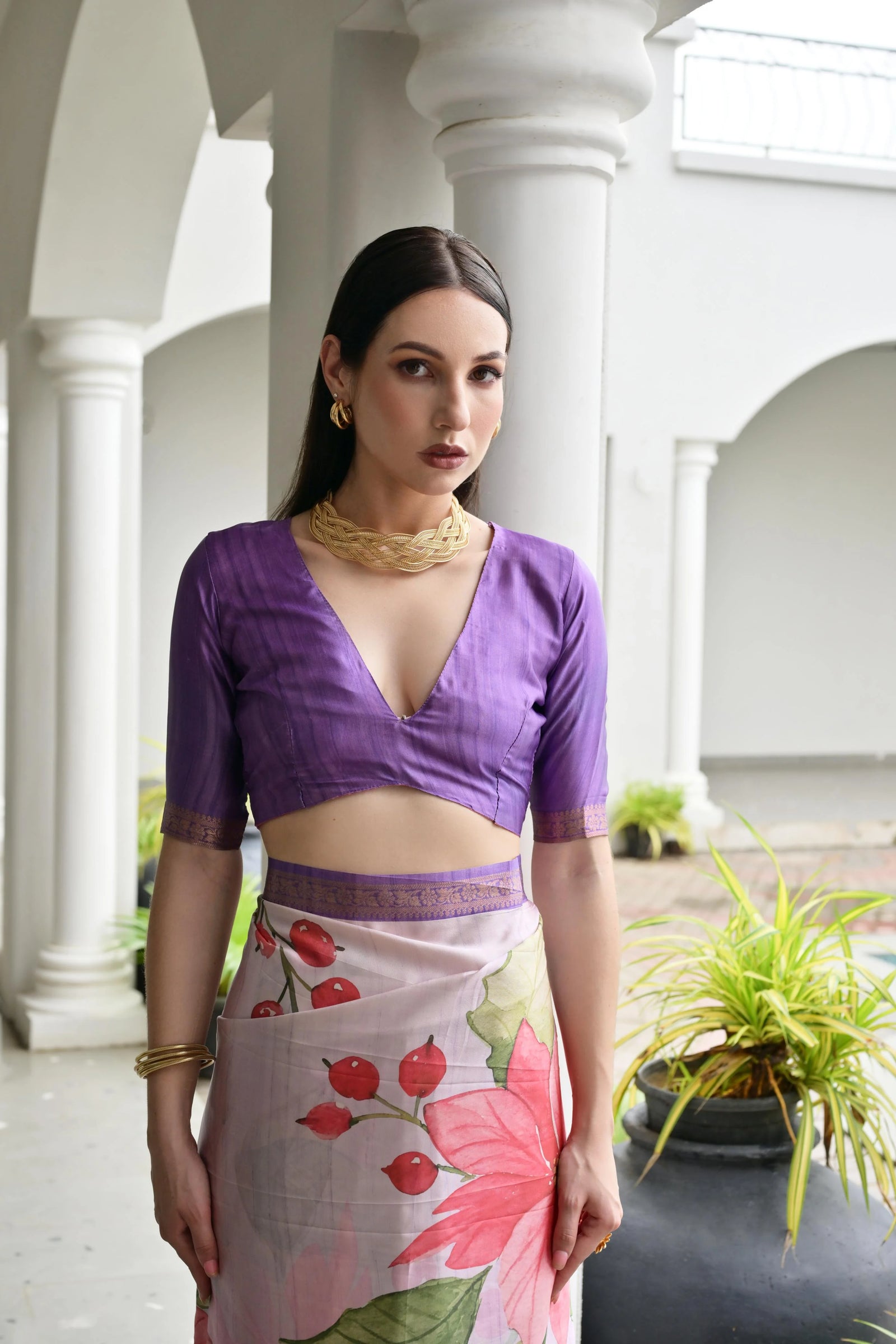 Woman wearing a purple top and floral skirt in an outdoor setting with columns and plants.