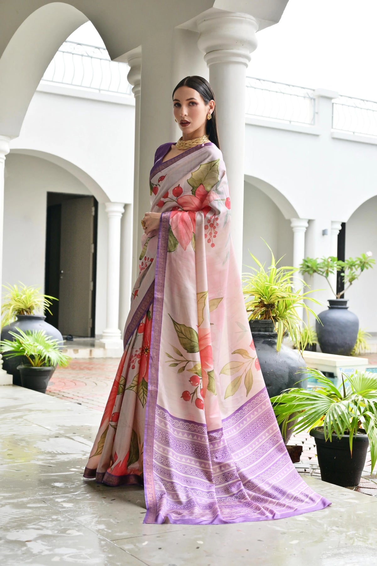 Woman in a colorful saree standing in an outdoor setting with plants and architectural columns.