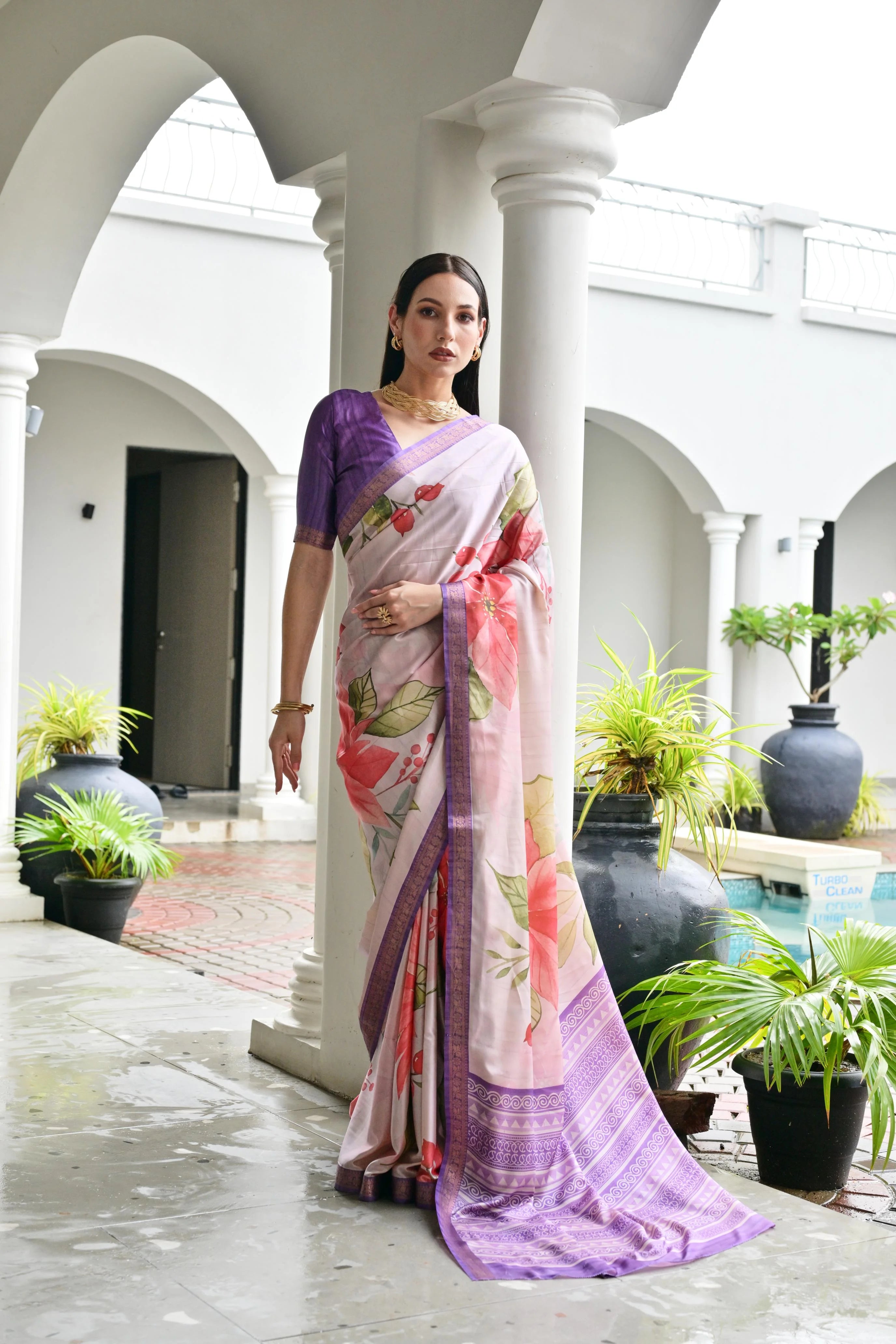 Woman in a floral saree standing in an outdoor setting with white columns and plants.