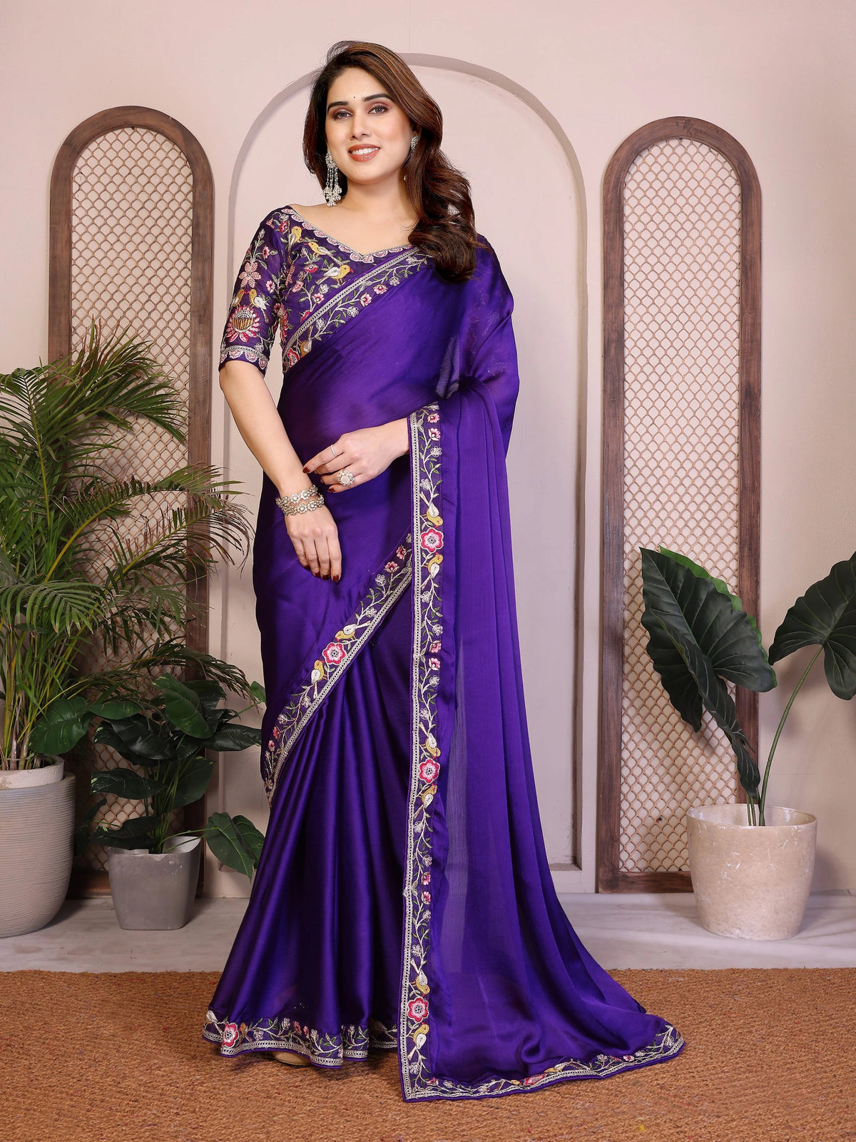 Woman wearing a purple saree with floral patterns in an indoor setting with plants.