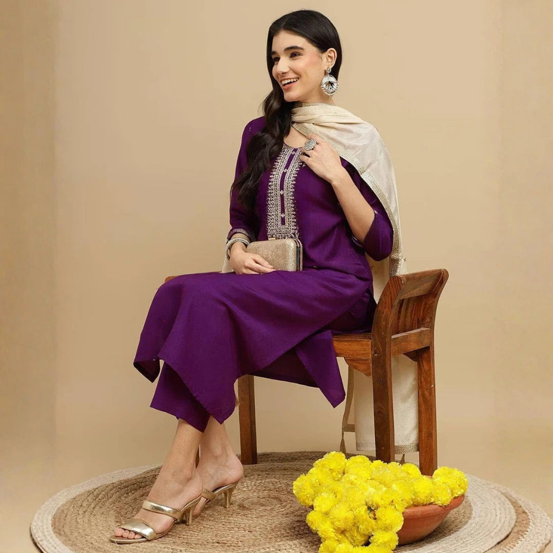 Woman in a purple outfit with a white scarf sitting on a wooden chair against a beige background.