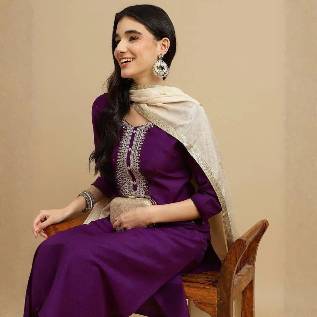 Woman in a purple outfit with a beige scarf sitting on a wooden chair against a beige background