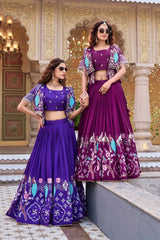 Two women pose in a traditional courtyard. One wears a vibrant indigo purple lehenga and the other a deep magenta version. Both outfits feature pleated silk skirts with elaborate peacock and floral embroidery along the hem, paired with a matching crop top and a short, open-front jacket with pearl-drop fringe.