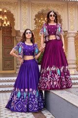 A full-length shot of the two women standing near a stone ledge. The image highlights the intricate turquoise and pink threadwork on the jackets and the heavy, traditional motifs—including swastikas and lotuses—embroidered on the lower half of the skirts.