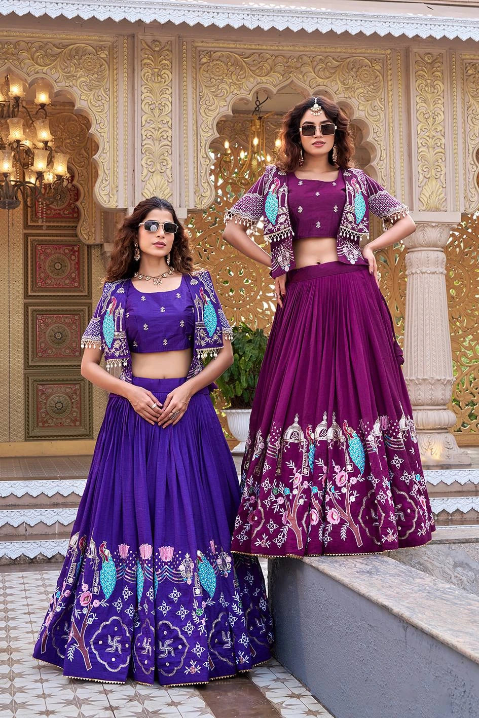 A front-facing pose of the duo standing under ornate golden arches. Both women wear modern square sunglasses, creating a contemporary fusion look with their traditional peacock-themed silk attire.
