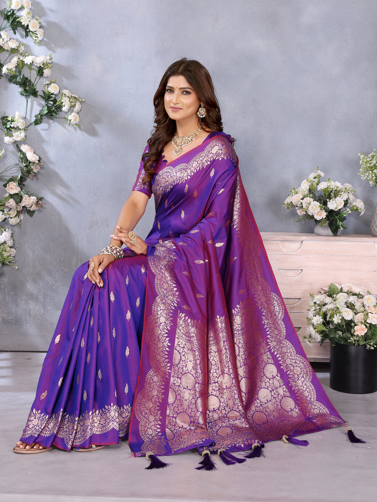 Woman in a purple saree with gold patterns sitting on a bench with floral decorations.