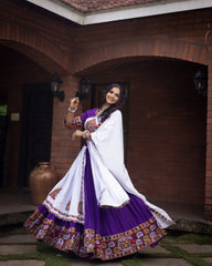 Woman in a traditional outfit with a white and purple saree standing in front of a brick wall.