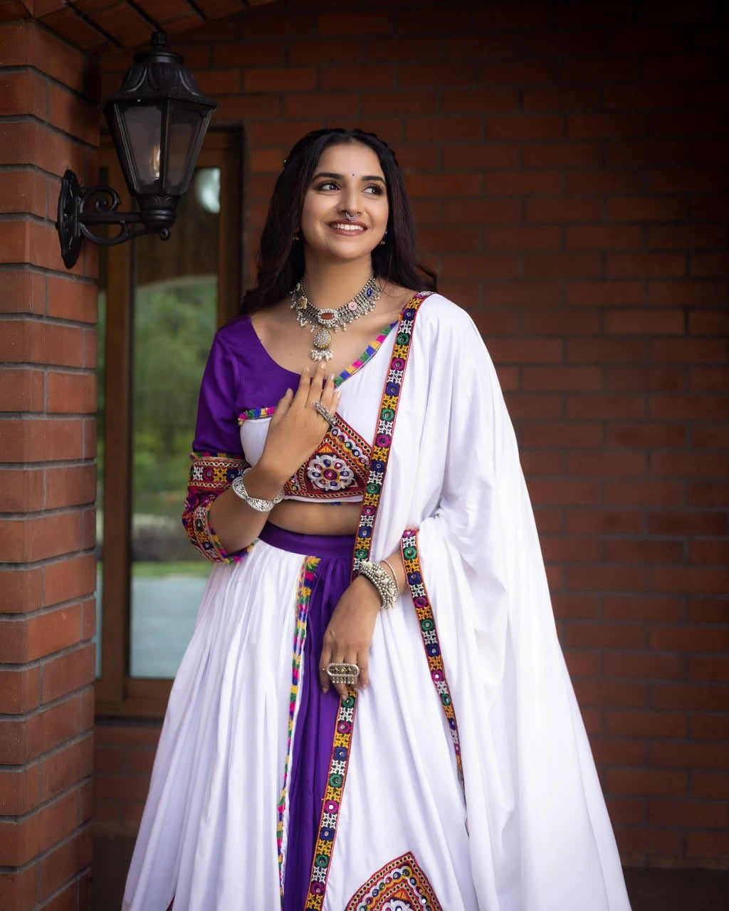 Woman in a traditional outfit with a purple blouse and white saree standing against a brick wall.