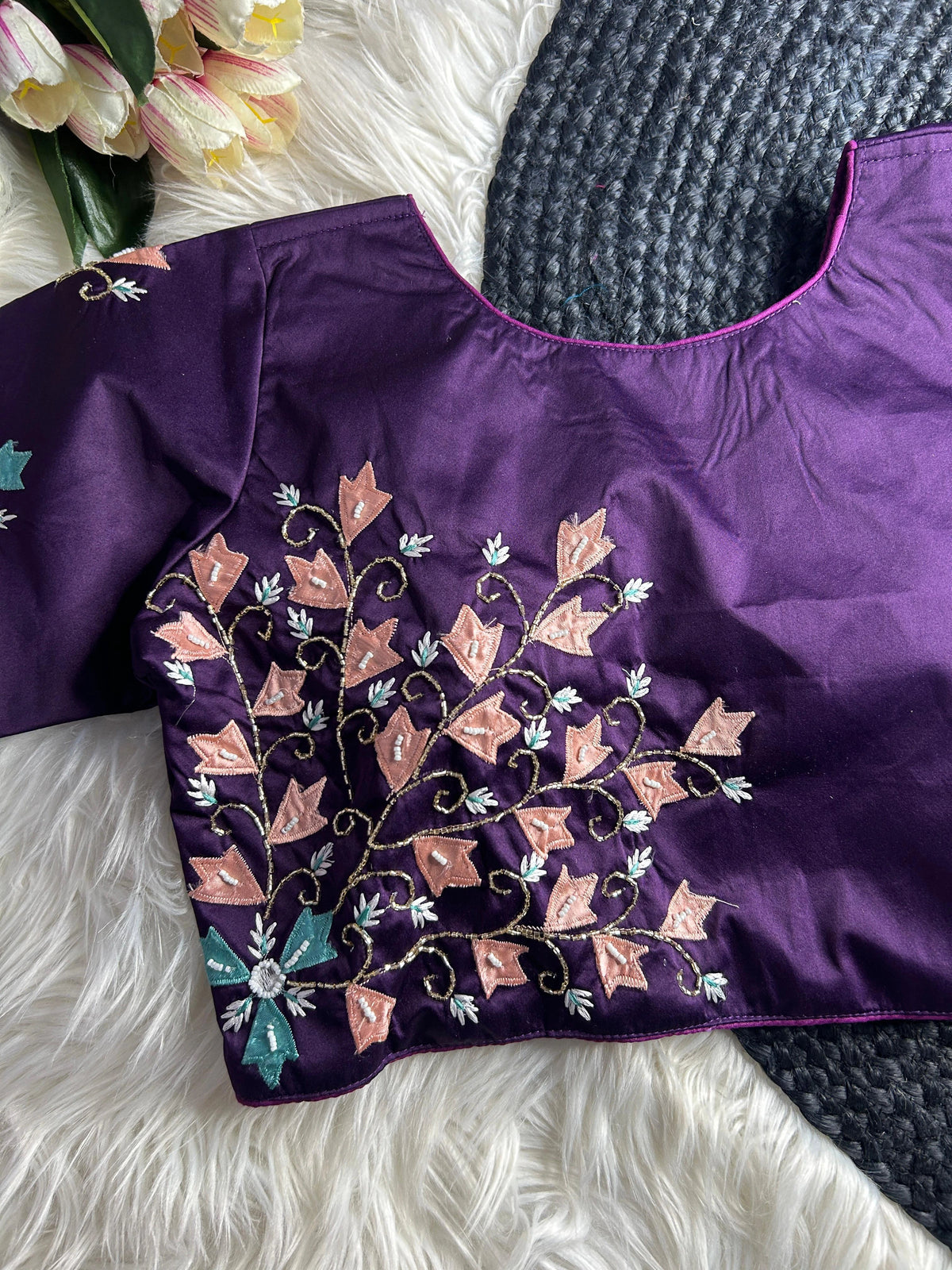 A deep purple silk saree blouse with a round neckline, featuring intricate hand-embroidery on the front left side. The design showcases winding gold-toned vines adorned with peach-colored leaves, white beaded accents, and a teal floral motif with a mirrored center at the bottom. The blouse is laid flat on a white faux-fur surface next to a grey woven mat and a bouquet of pink and white tulips.