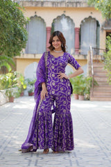 Woman in a purple traditional outfit standing outdoors with a building and greenery in the background