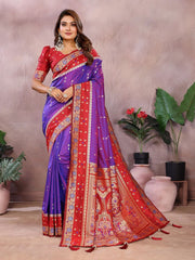Woman wearing a traditional purple and red saree with a decorative border, standing against a neutral background with plants.