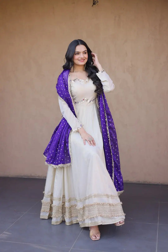 Woman in a white traditional outfit with a purple dupatta against a beige wall.