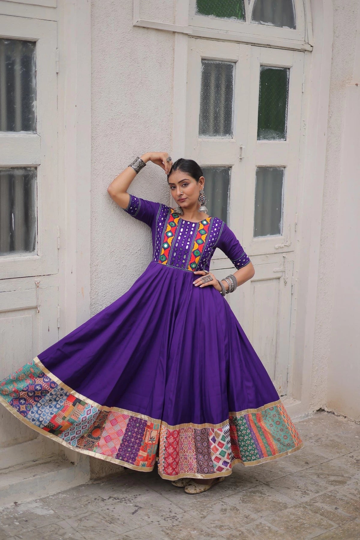 Woman in a vibrant purple traditional outfit with colorful patterns standing against a white wall.