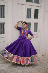 Woman in a vibrant purple traditional outfit with colorful patterns standing against a white wall.