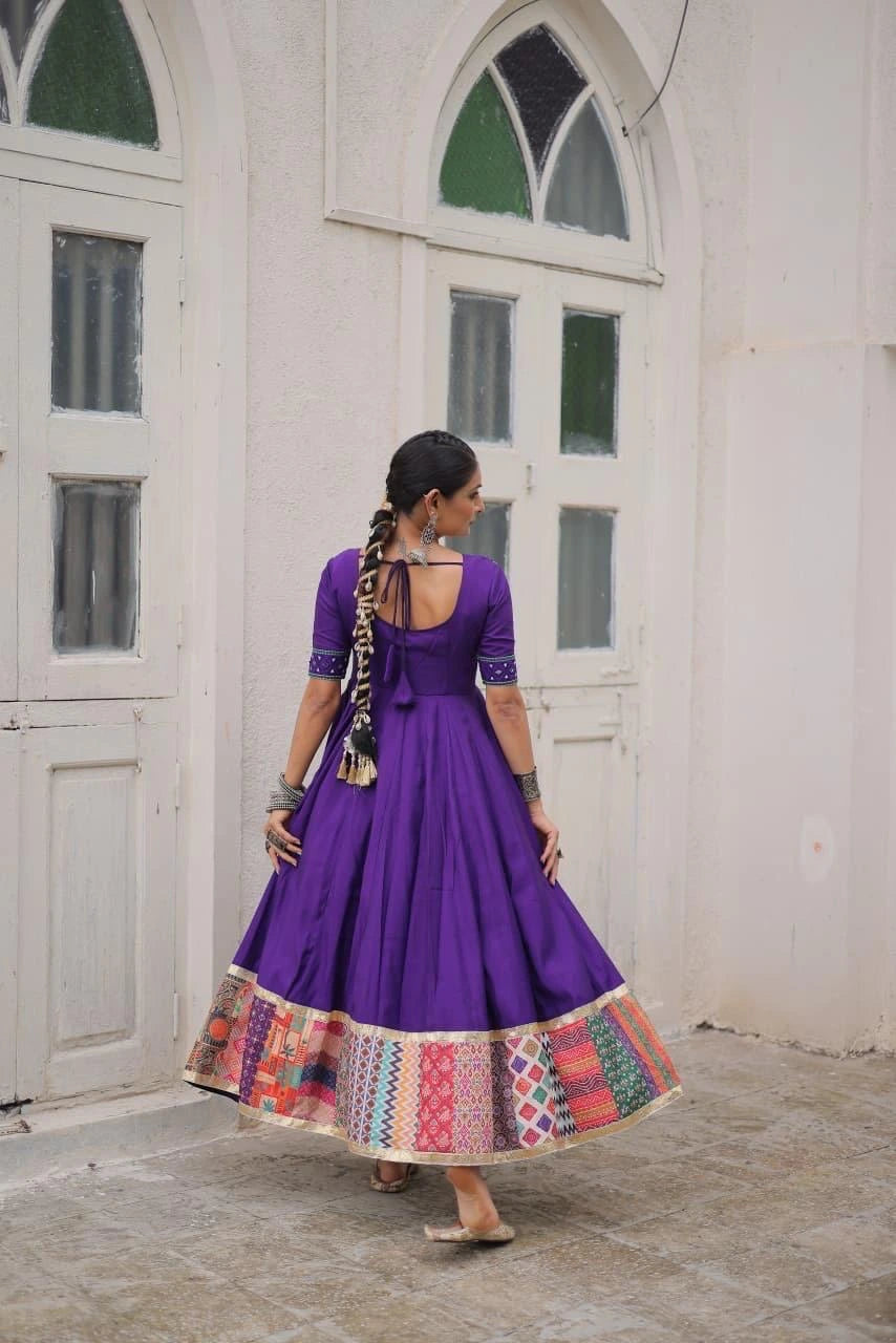 Woman in a purple dress with a colorful skirt standing in front of a white building.