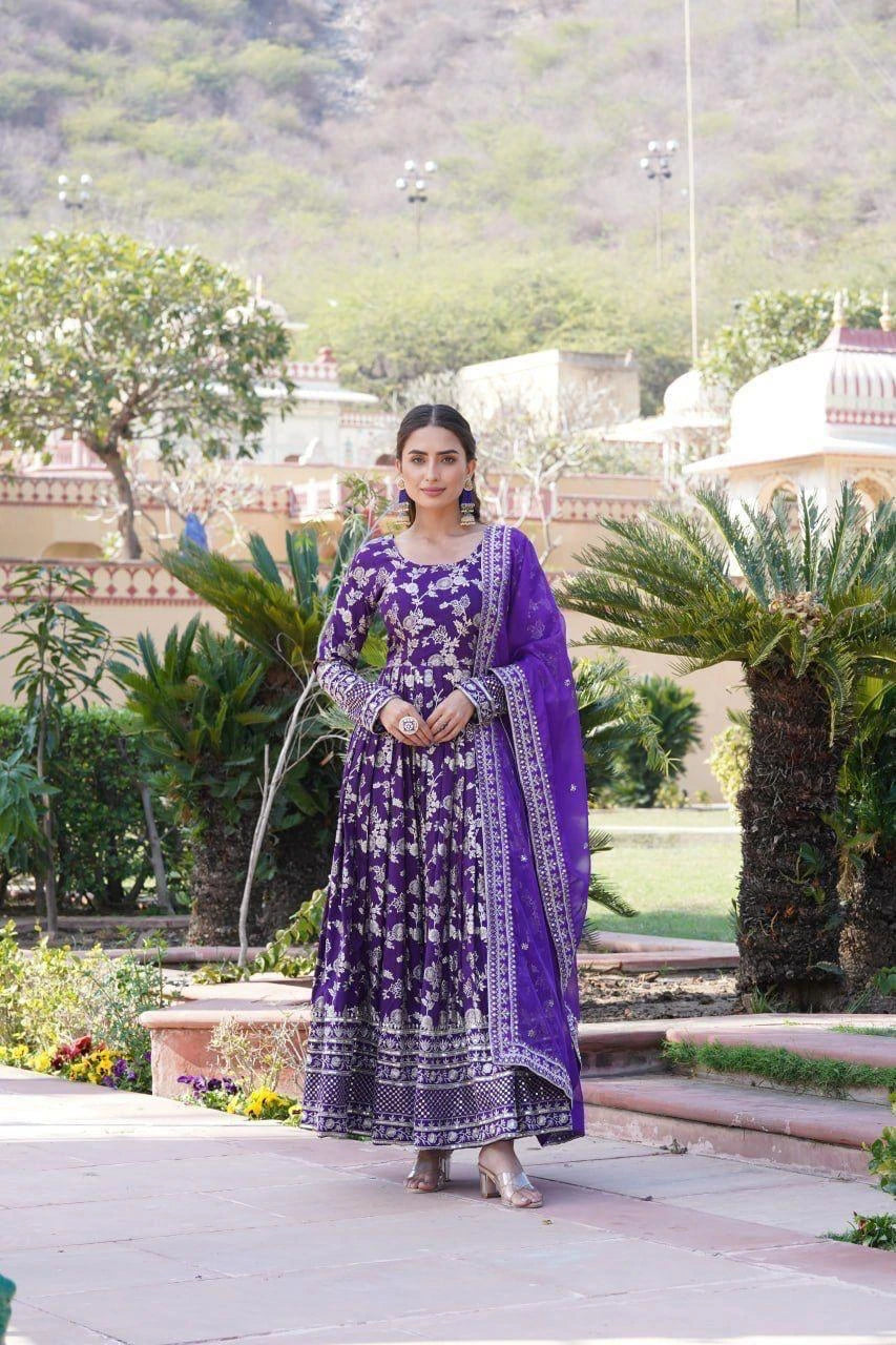 Woman in a purple traditional outfit standing in a garden with greenery and architectural elements in the background.