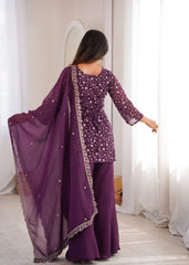 Woman wearing a purple traditional outfit with a sheer dupatta in a room with white curtains.