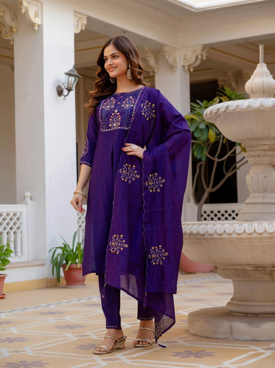 Woman in a purple traditional outfit standing in an outdoor setting with decorative columns and plants.