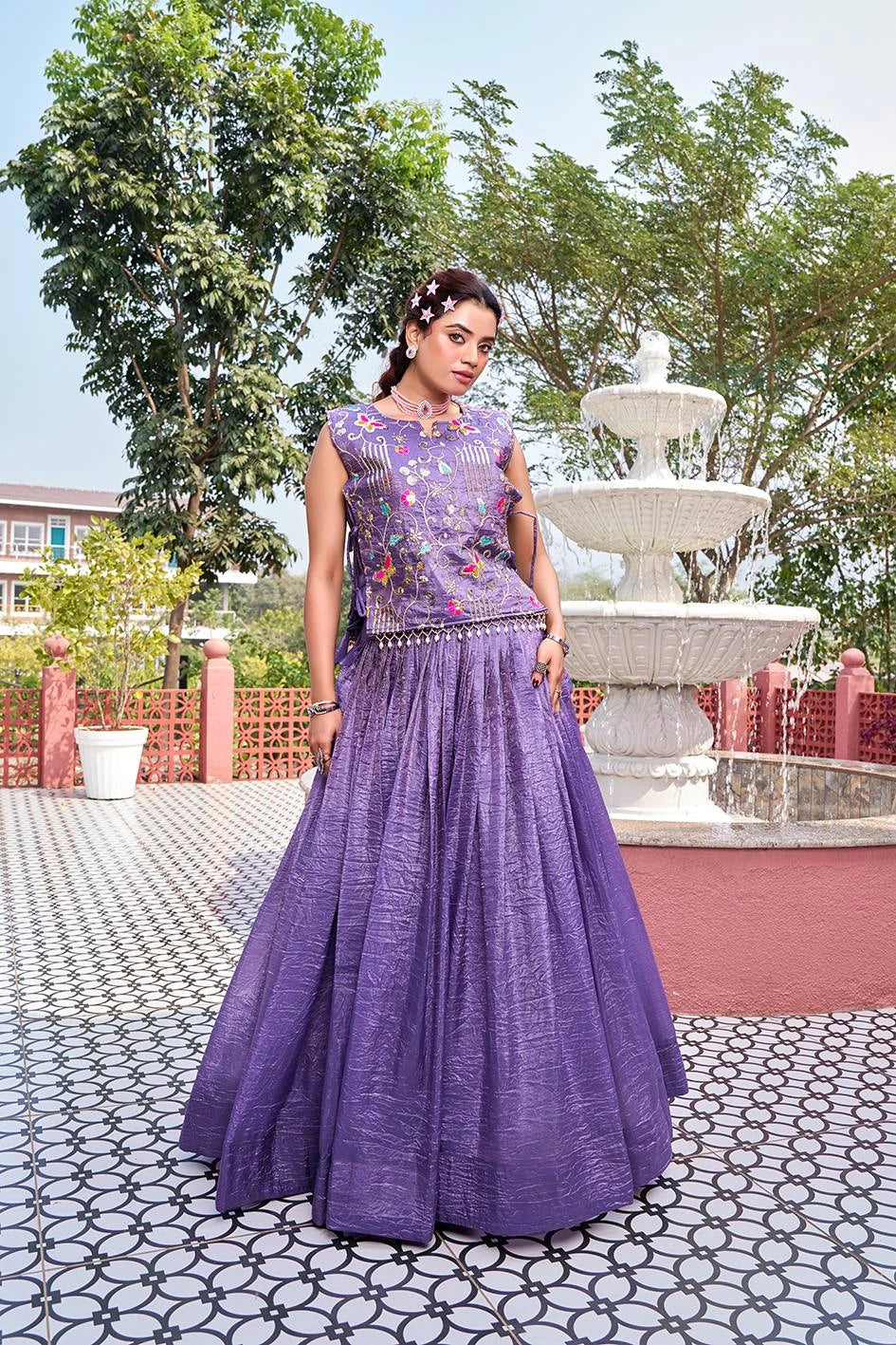 Woman in a purple traditional outfit standing in front of a fountain with trees in the background
