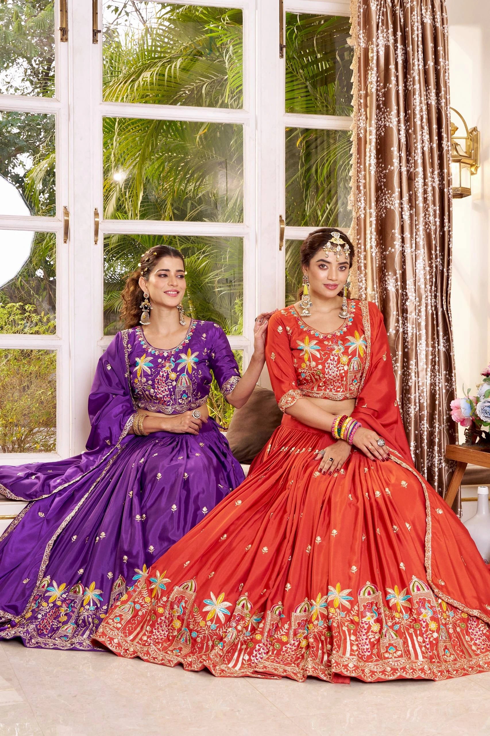 Two women in traditional embroidered lehengas sitting indoors with a window and curtain in the background.