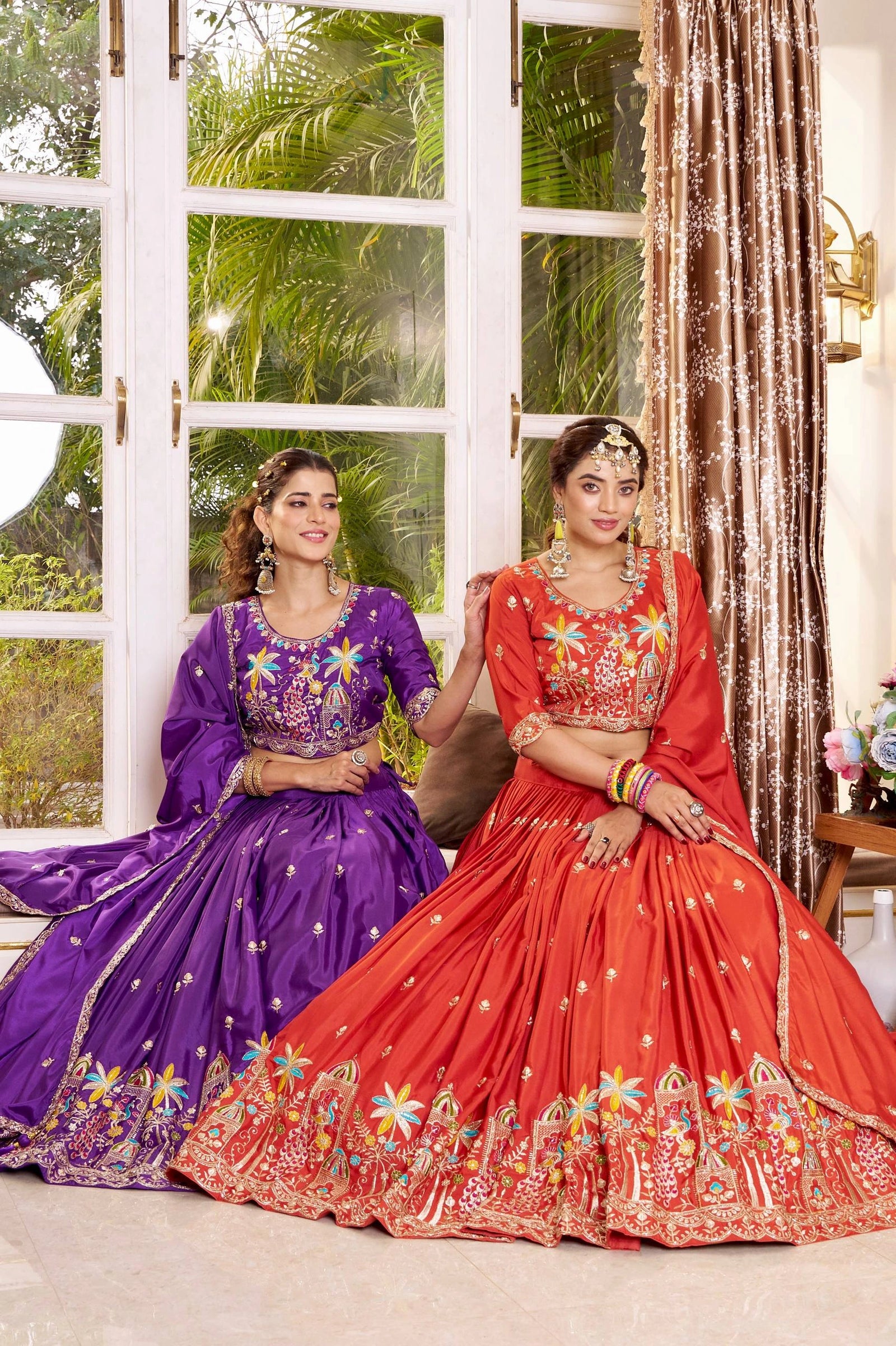 Two women in traditional embroidered lehengas sitting indoors with a window and curtain in the background.