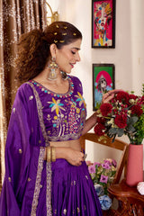 Woman in a purple traditional outfit with floral patterns, standing in a room with decorative elements.