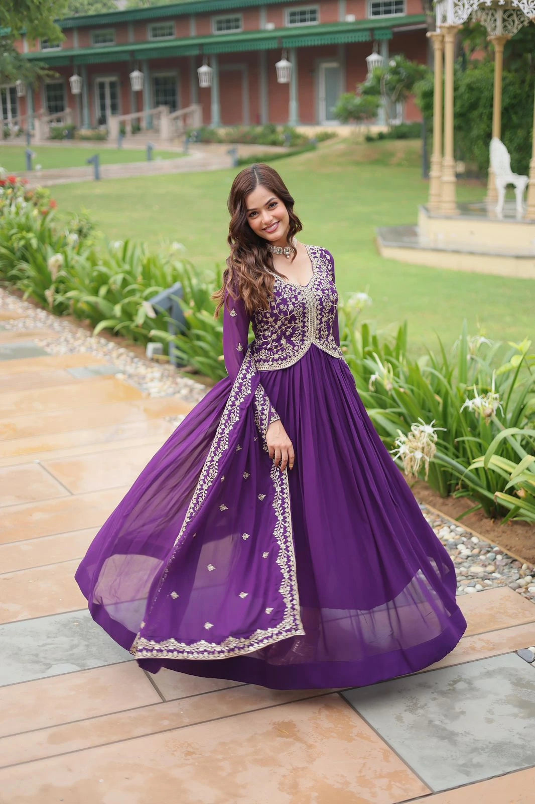 Woman in a purple gown standing outdoors on a stone path with greenery and a building in the background.