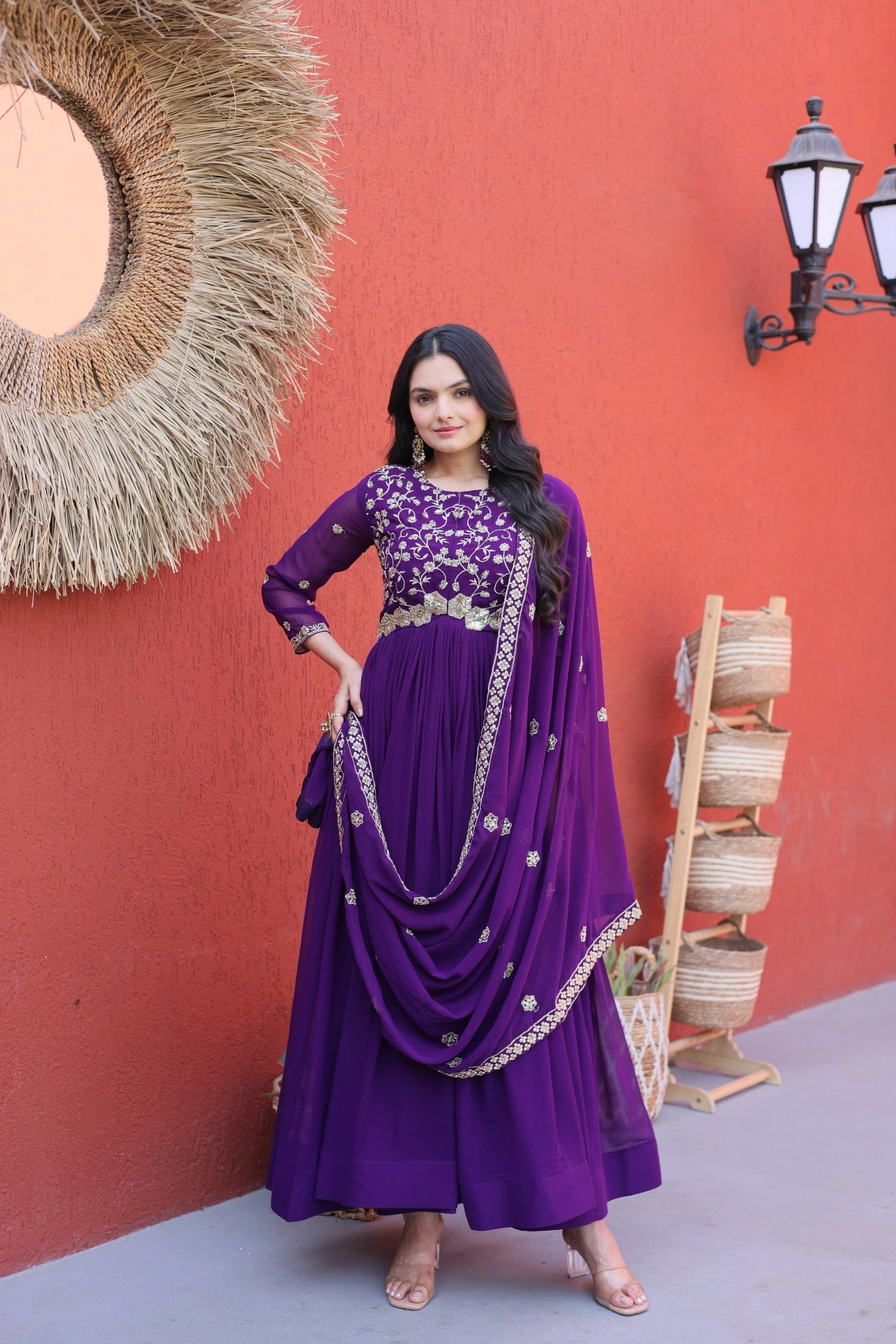 Woman in a purple traditional outfit standing against a red wall with decorative elements.