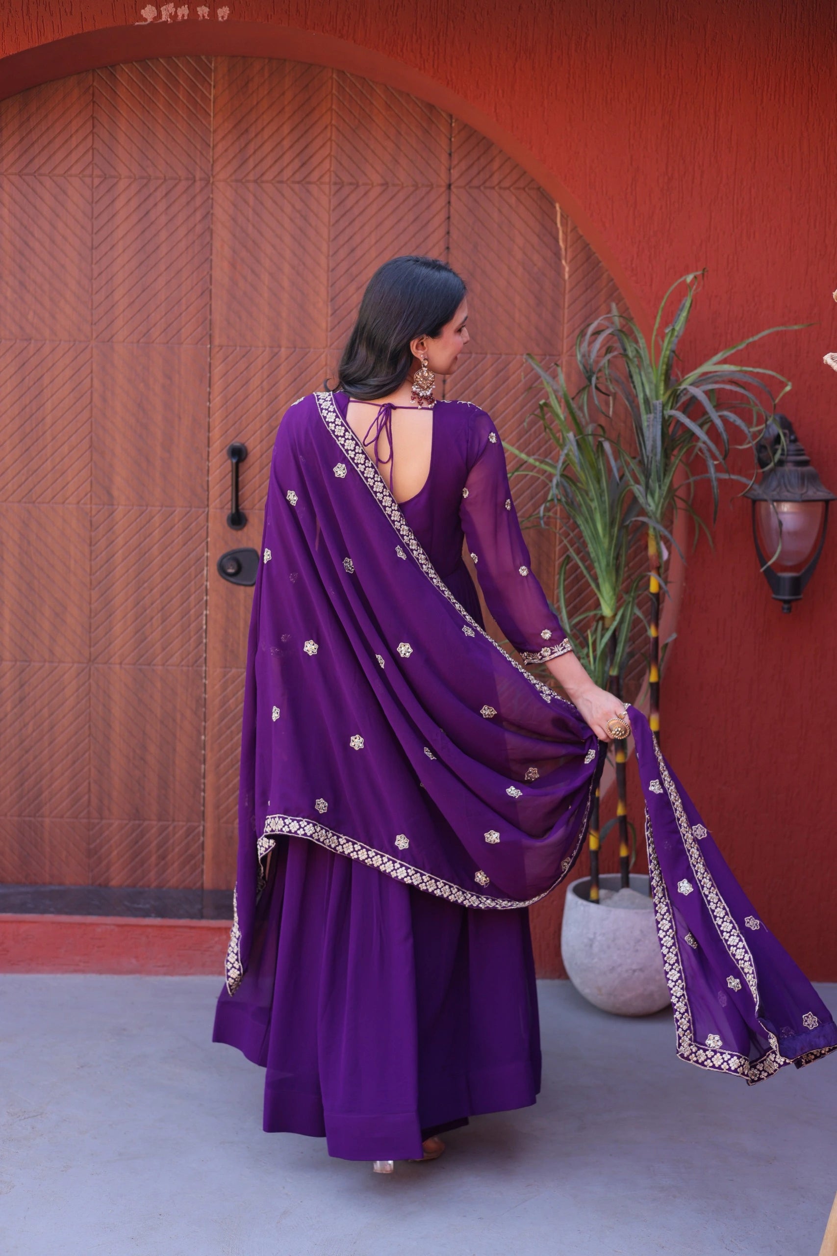 Woman in a purple traditional outfit standing in front of a red wall with a plant.