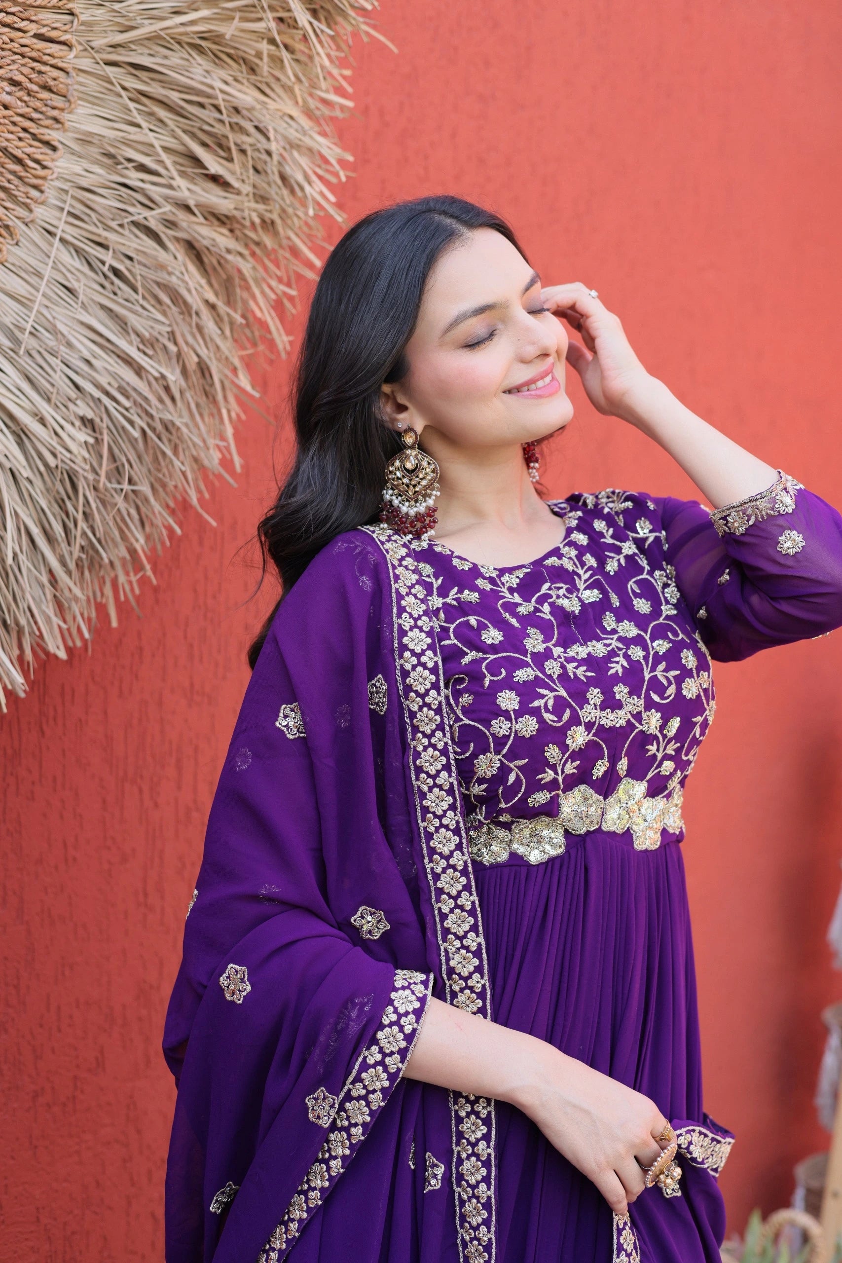 Woman wearing a purple traditional outfit with gold embroidery against a red wall.