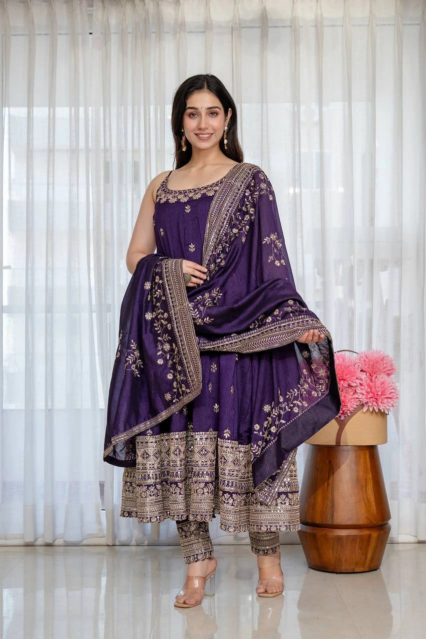 Woman in a traditional purple and gold outfit standing in front of a white curtain with pink flowers on a wooden stool.