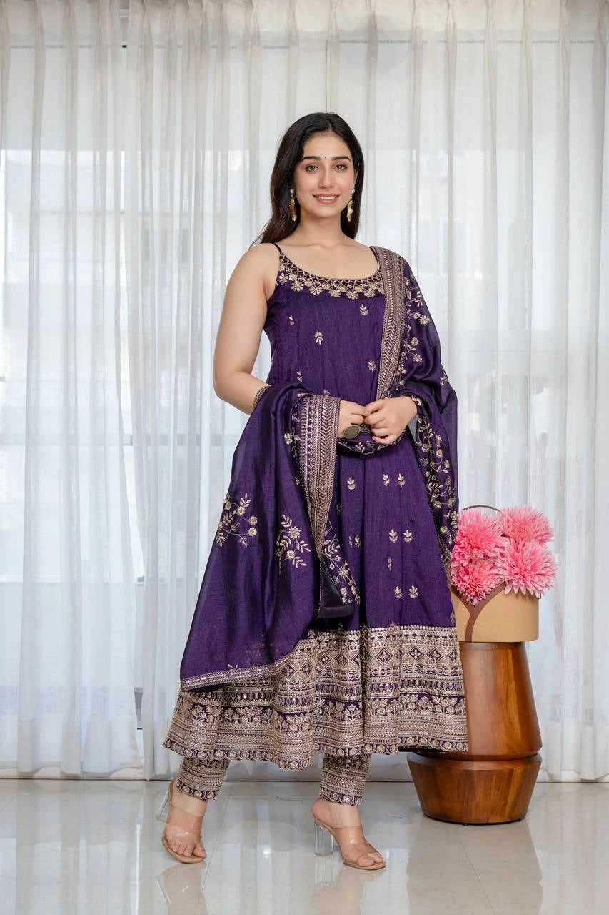 Woman in a purple and brown traditional outfit standing next to a wooden stool with pink flowers against a white curtain background.