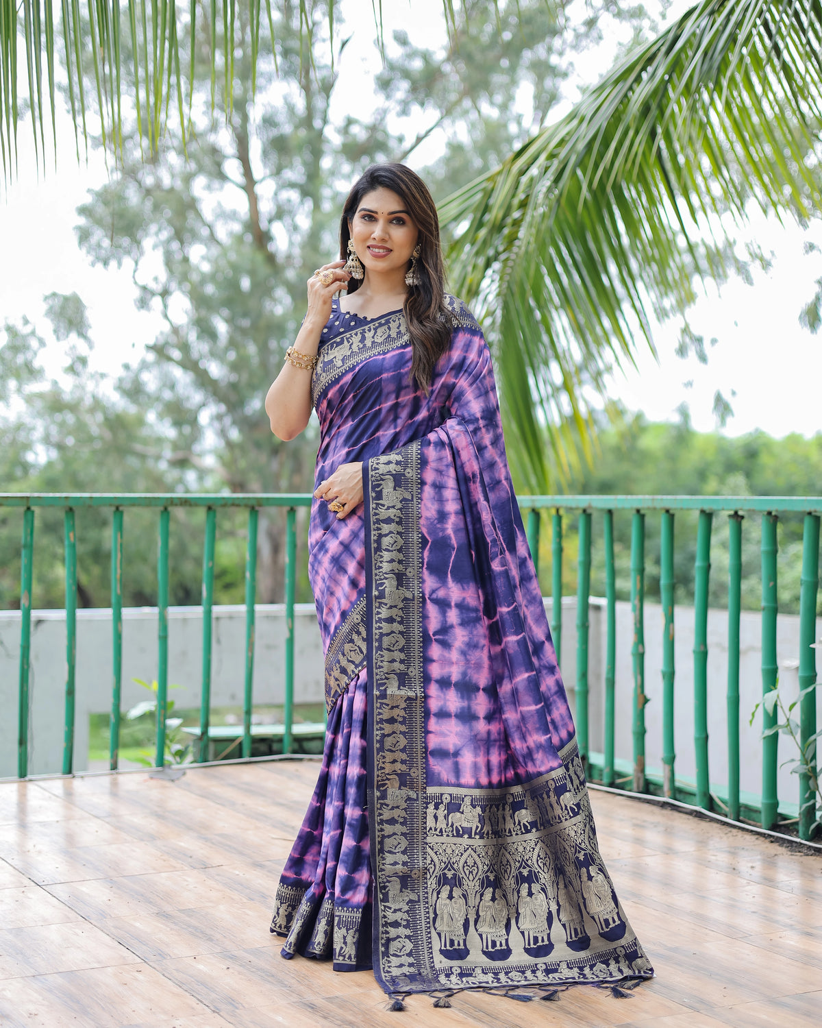 Woman in a purple and black saree standing on a wooden deck with greenery in the background